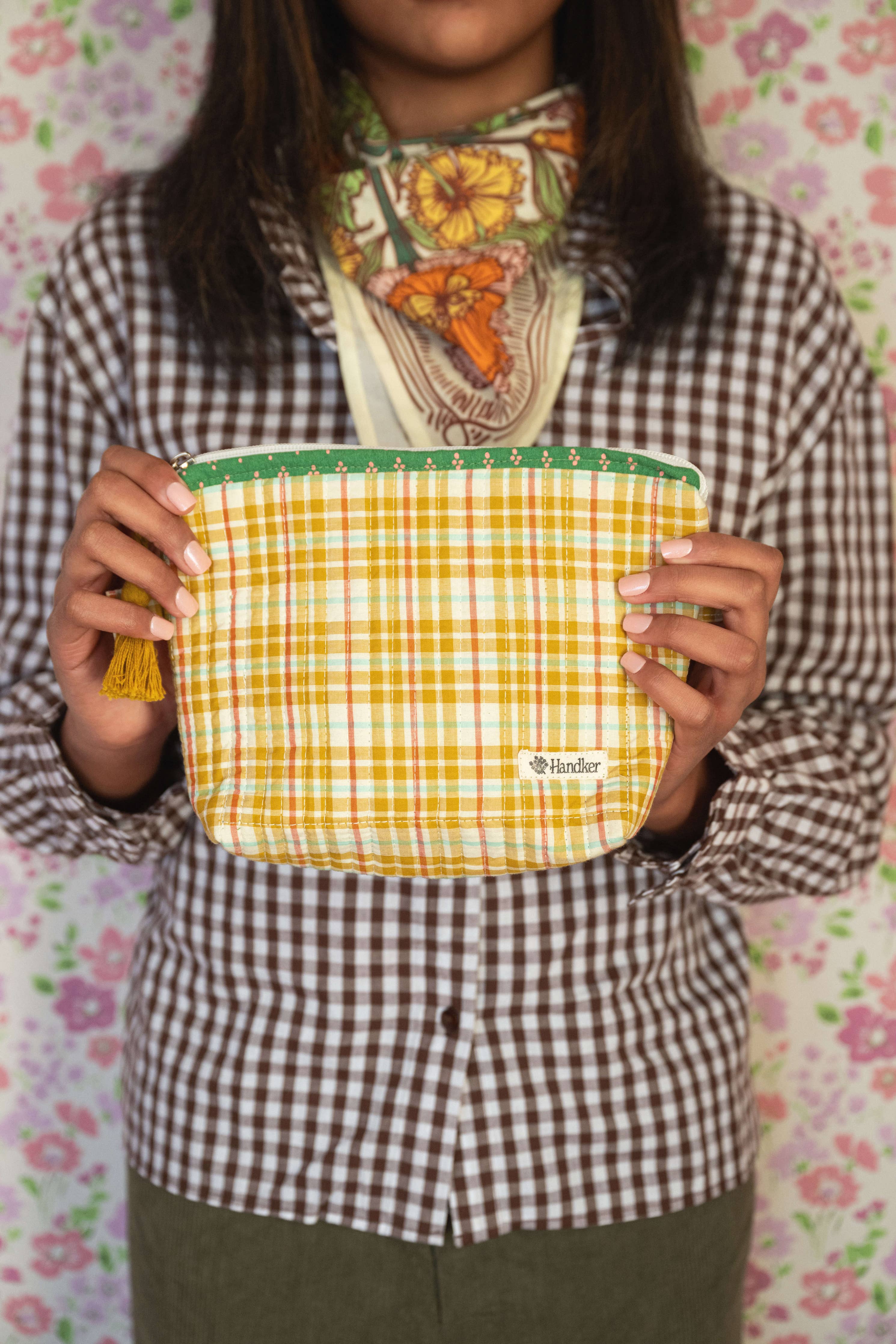 Person holding a yellow and green checkered pouch with a floral-patterned scarf around their neck against a floral background.