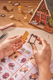 Person assembling a miniature model of a house on a grid-patterned surface with tools and materials around.