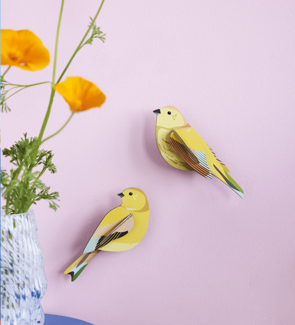 Two yellow bird-shaped decorations on a pink wall with a vase of flowers.