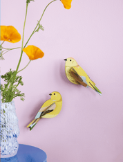 Two yellow bird-shaped decorations on a pink wall with a vase of flowers.