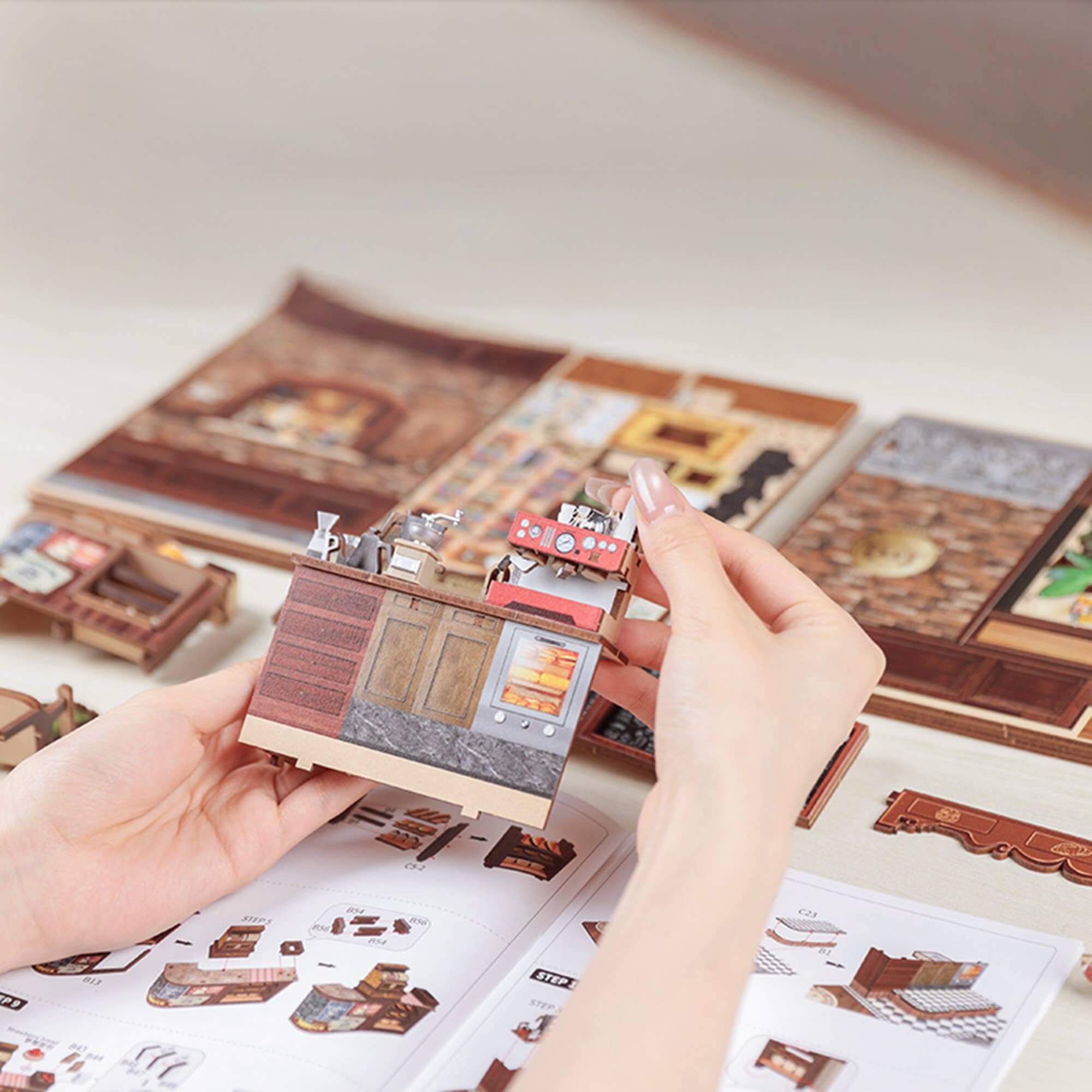 Hand holding a miniature model of a building with a detailed interior, against a background of similar models and design sheets.