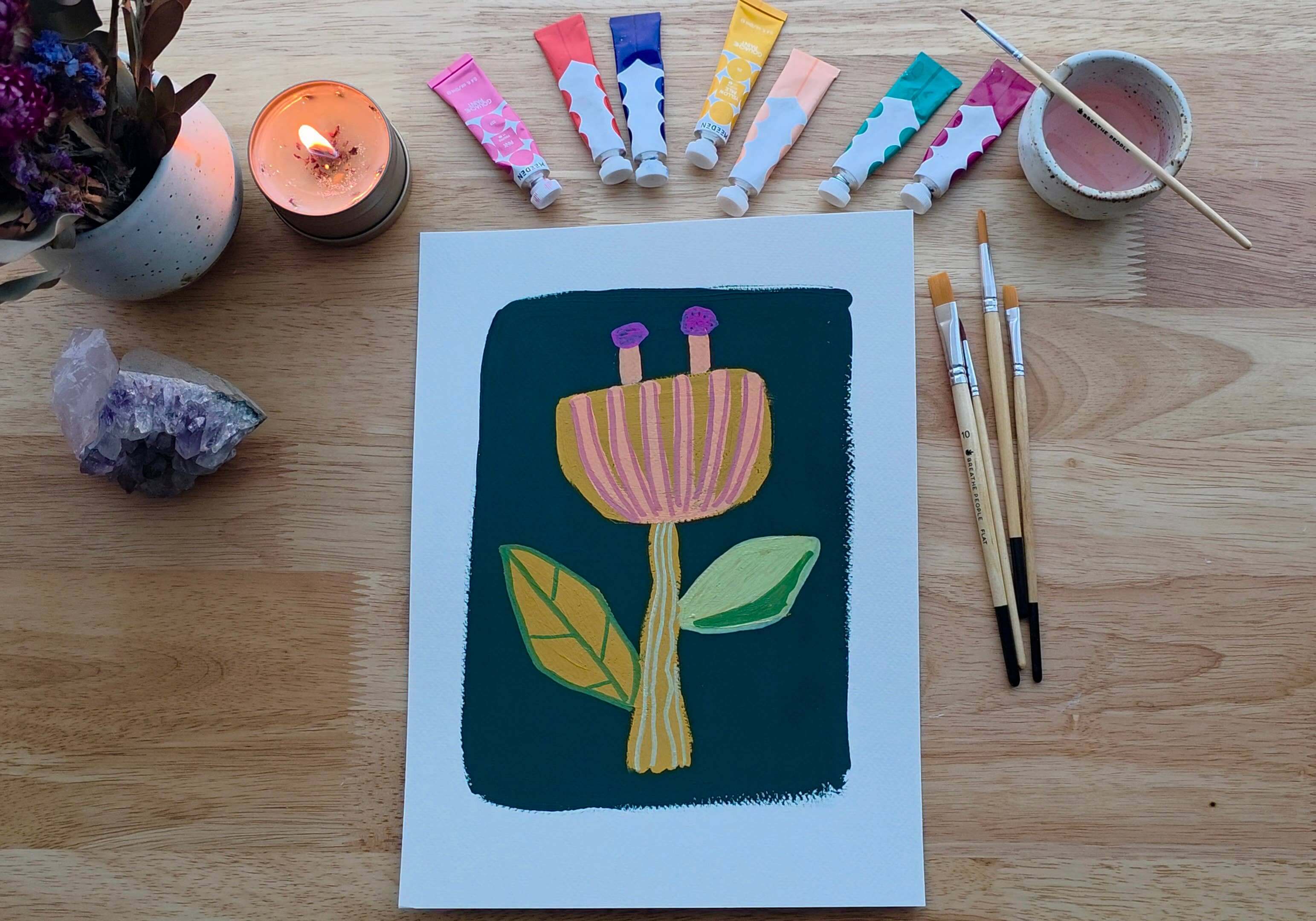 Artistic depiction of a flower with paint tubes, brushes, and a candle on a wooden surface.