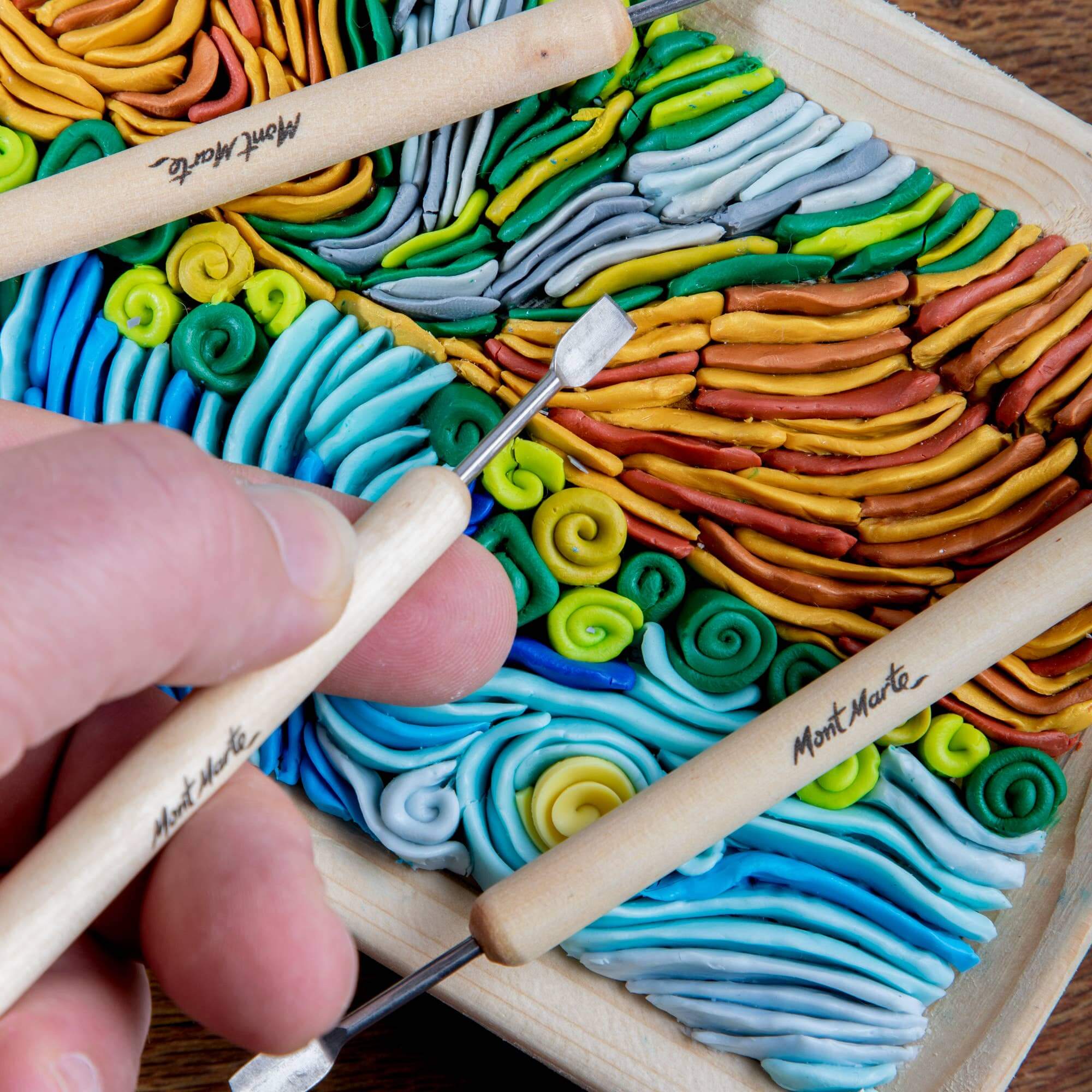 Colorful swirled clay with a hand holding small tools on a wooden board.