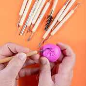 Hands using a small clay tool on pink clay with other tools in the background on an orange surface