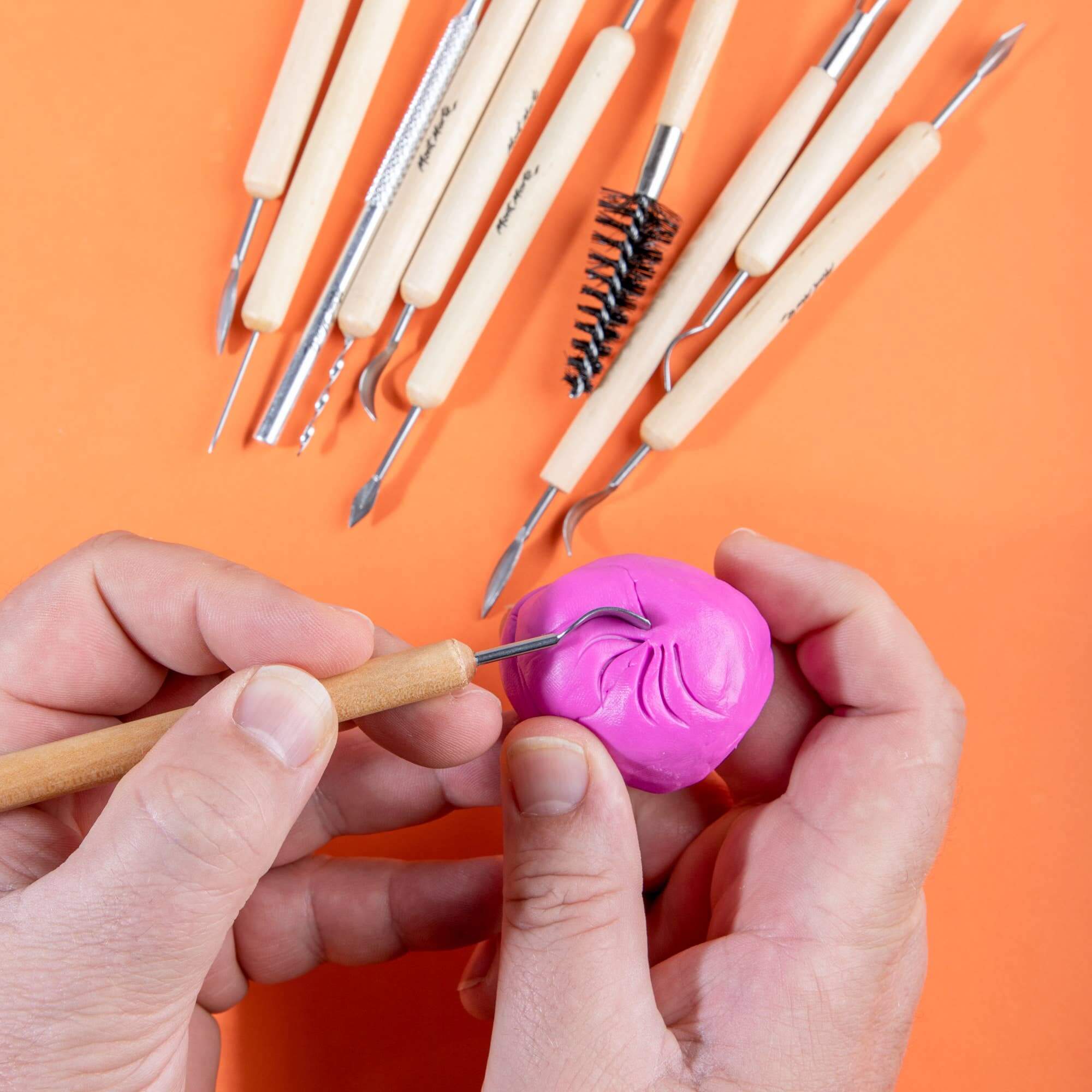 Hands using a small clay tool on pink clay with other tools in the background on an orange surface