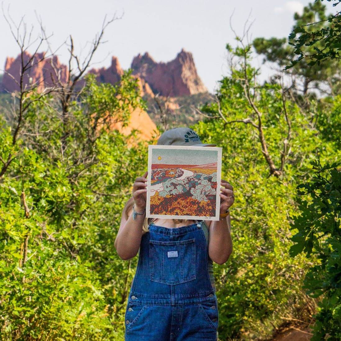 Person holding a framed landscape print in front of their face with a scenic background of trees and mountains.