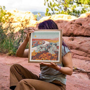 Person holding a framed artwork in front of their face with a natural landscape background