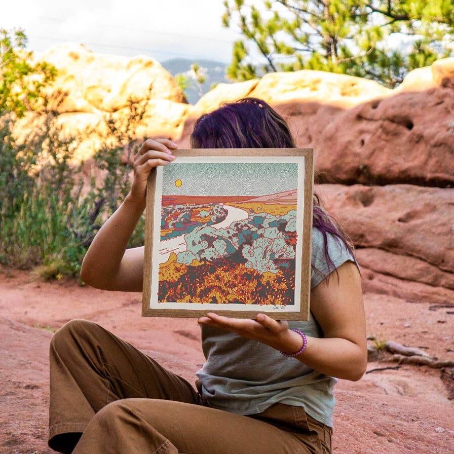Person holding a framed artwork in front of their face with a natural landscape background