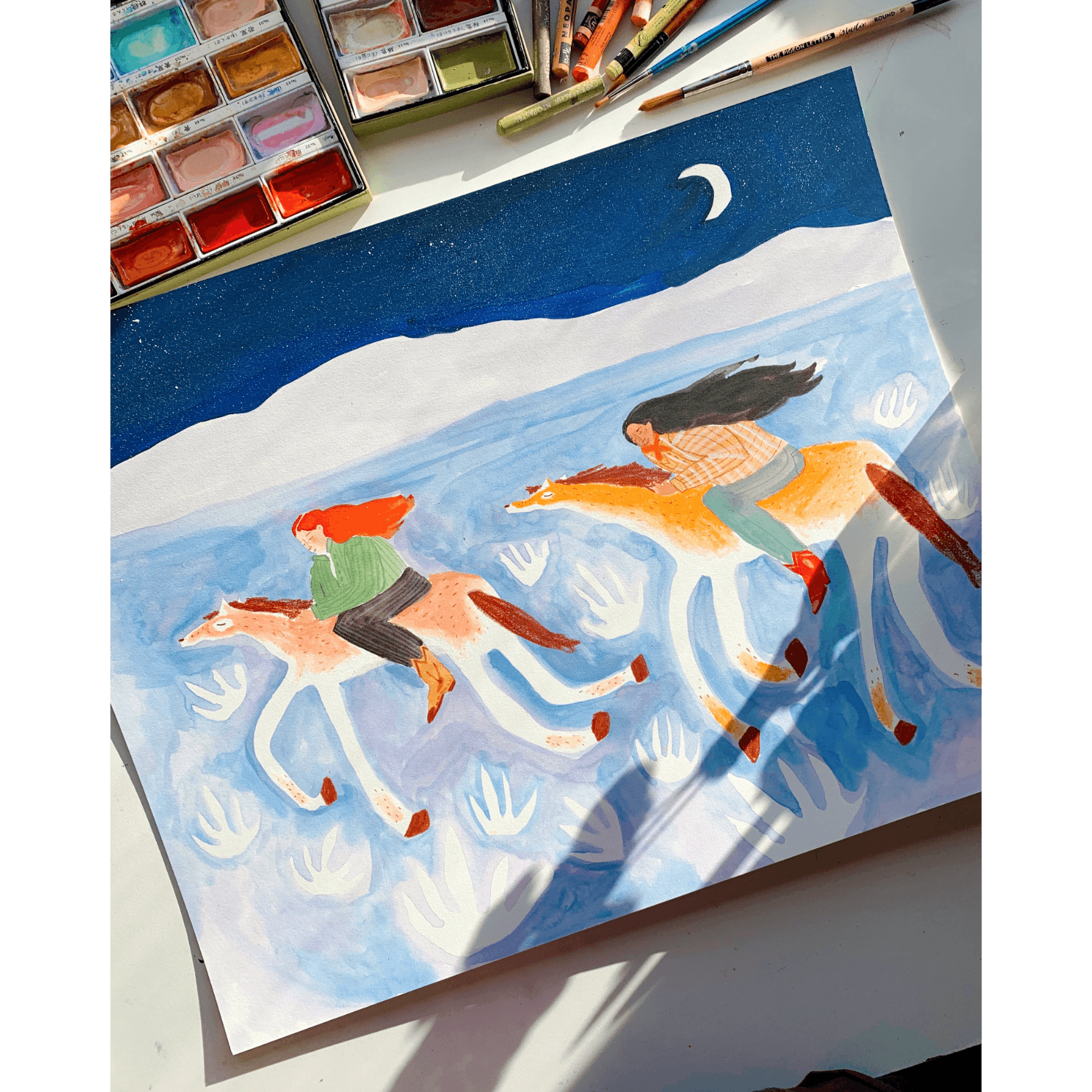Painting of two cowgirls on horseback with a watercolor palette lying on a desk and brushes in the background.