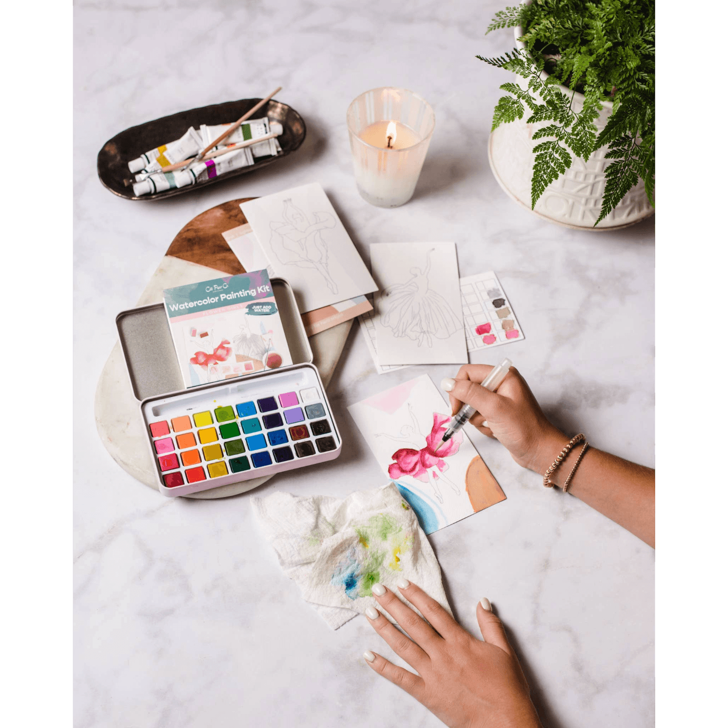 Person painting with watercolors on a marble surface with art supplies, a candle and plant.