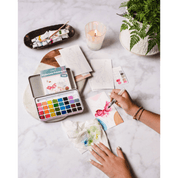 Person painting with watercolors on a marble surface with art supplies, a candle and plant.