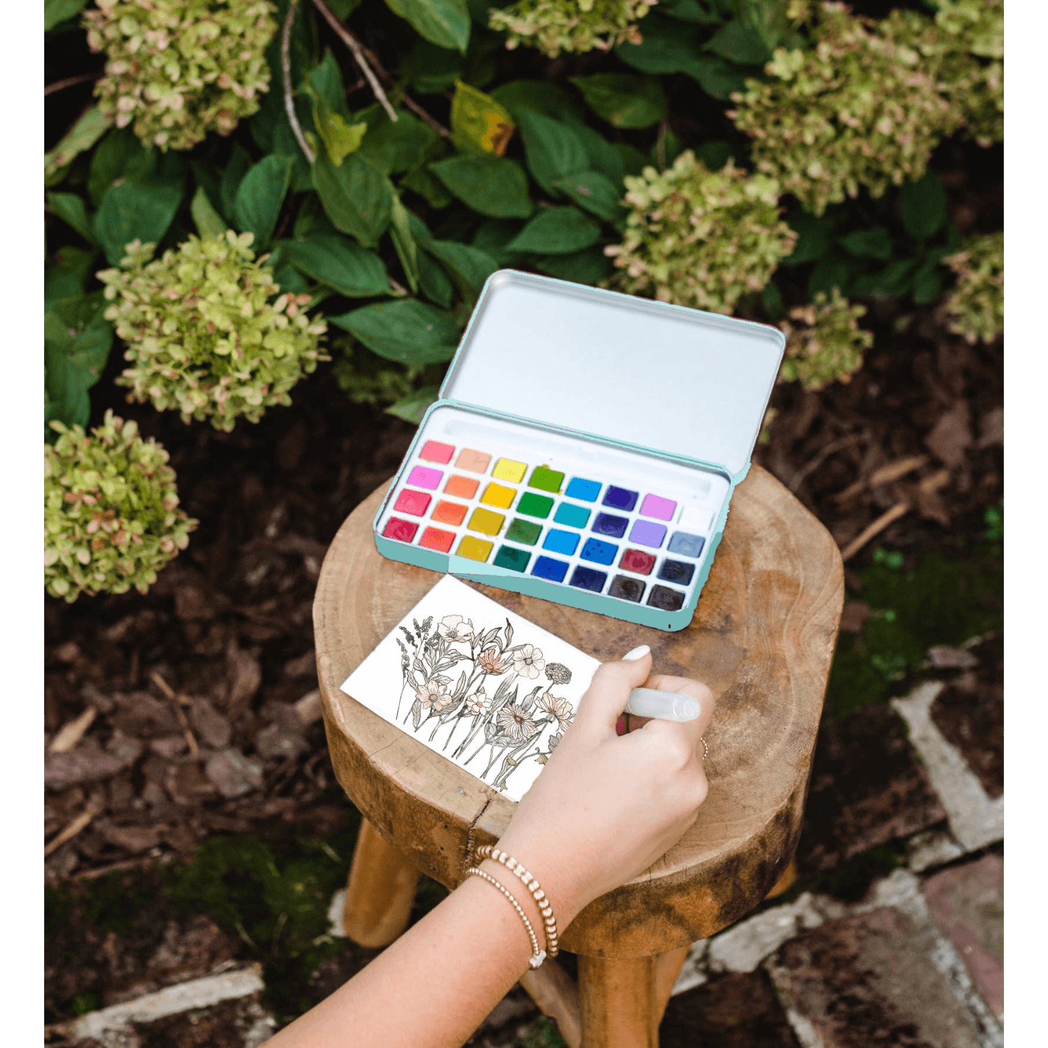 Person with watercolor set and paper outdoors, surrounded by greenery