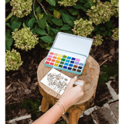Person with watercolor set and paper outdoors, surrounded by greenery