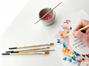 Person painting a floral design on paper with brushes and a small container of paint.