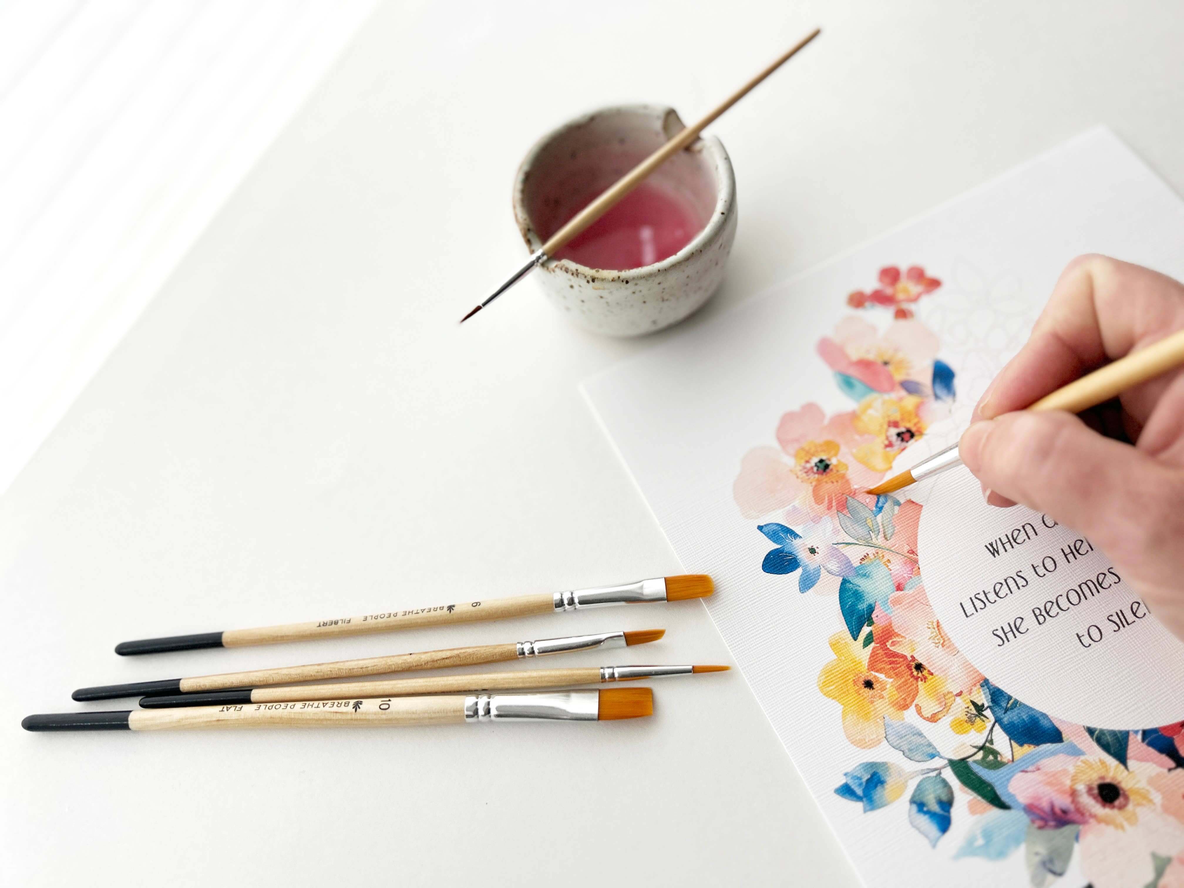 Person painting a floral design on paper with brushes and a small container of paint.