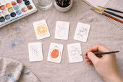 Artistic watercolor practice with small cards and a hand holding a brush on a cream linen cloth.