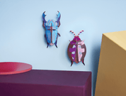 Colorful beetle-shaped paper figurines on a blue wall with a pink and yellow geometric object.