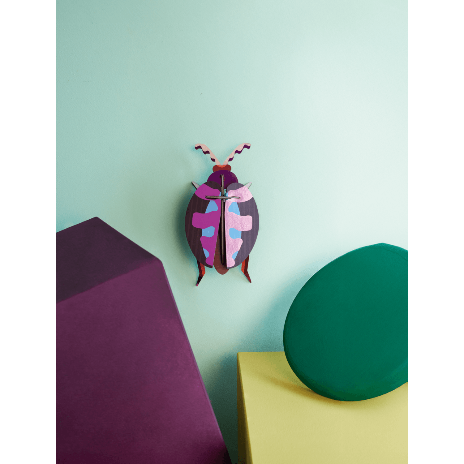 Colorful pink beetle shaped paper figurine on a light green wall with purple and green objects.