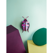 Colorful pink beetle shaped paper figurine on a light green wall with purple and green objects.