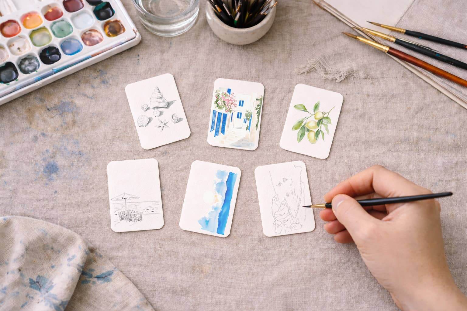 Hand painting small watercolor artworks on cards with a watercolor palette and brushes on a cream linen tablecloth.