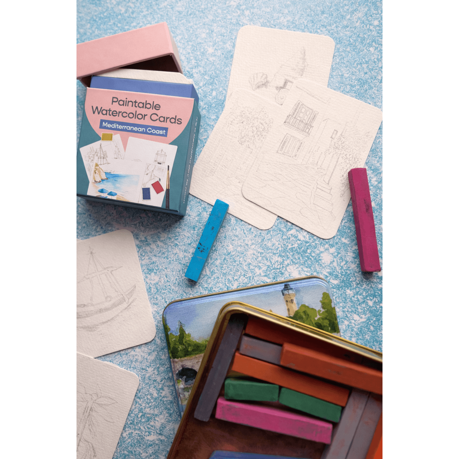 Paintable watercolor cards with a tin of paint sticks on a textured blue surface.