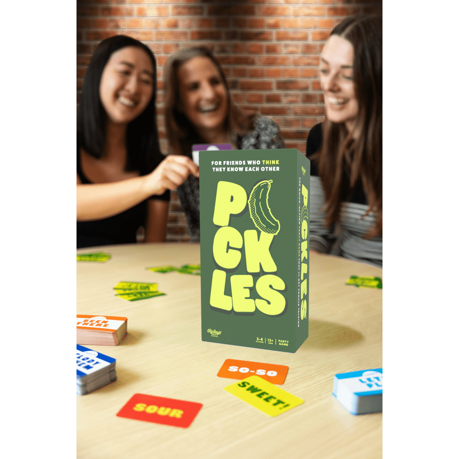 Board game packaging with 'PICKLES' text and banana illustration, surrounded by players and game pieces on a table.