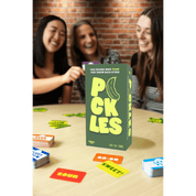 Board game packaging with 'PICKLES' text and banana illustration, surrounded by players and game pieces on a table.