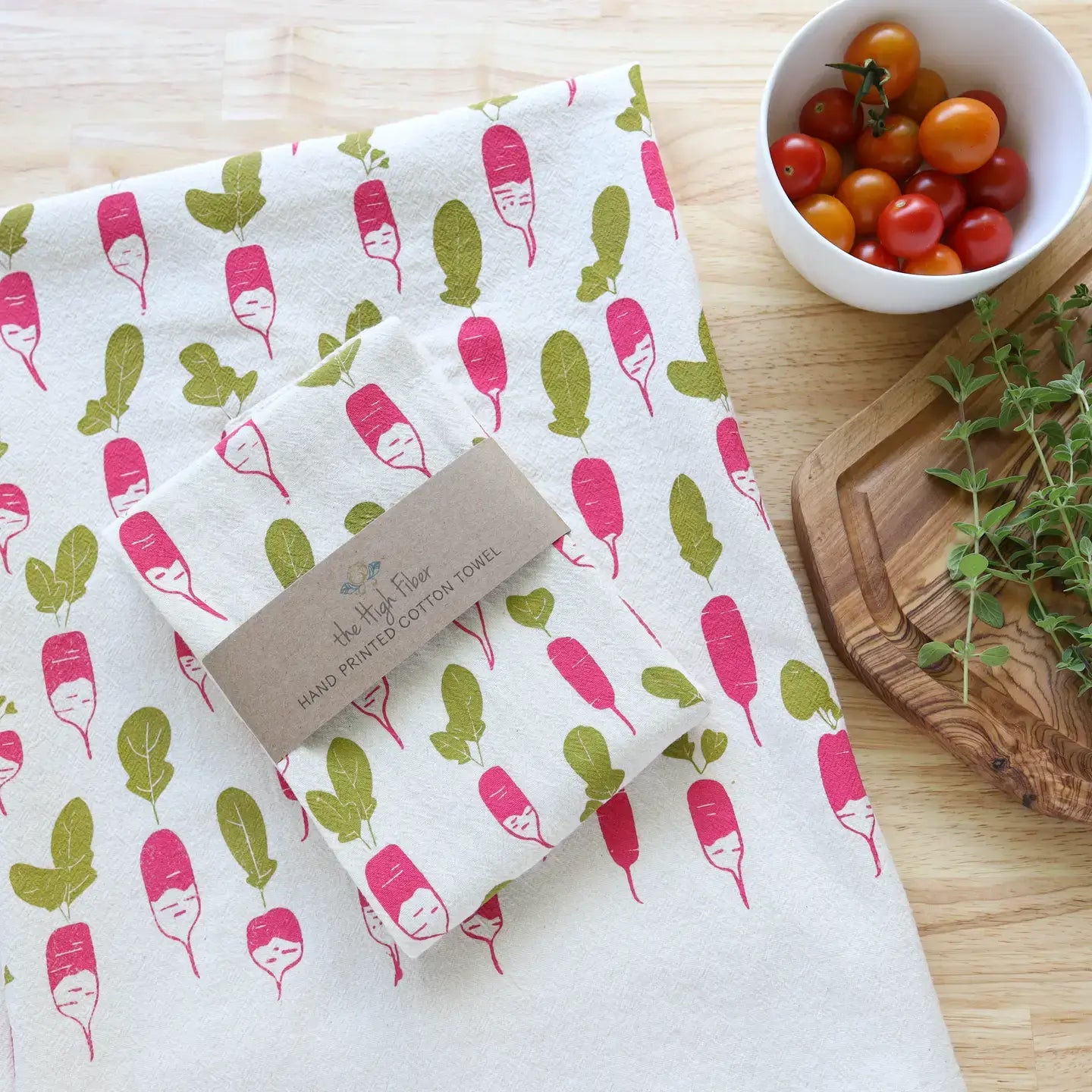 Folded fabric with radish pattern on a wooden surface with a bowl of tomatoes and a plant.