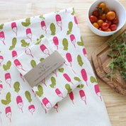 Folded fabric with radish pattern on a wooden surface with a bowl of tomatoes and a plant.