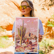 Woman holding a desert-themed print with cacti and a horse in a natural setting