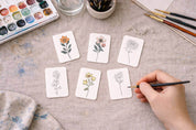 Hand painting watercolor illustrations of flowers on small cards with a palette and brushes in the background.
