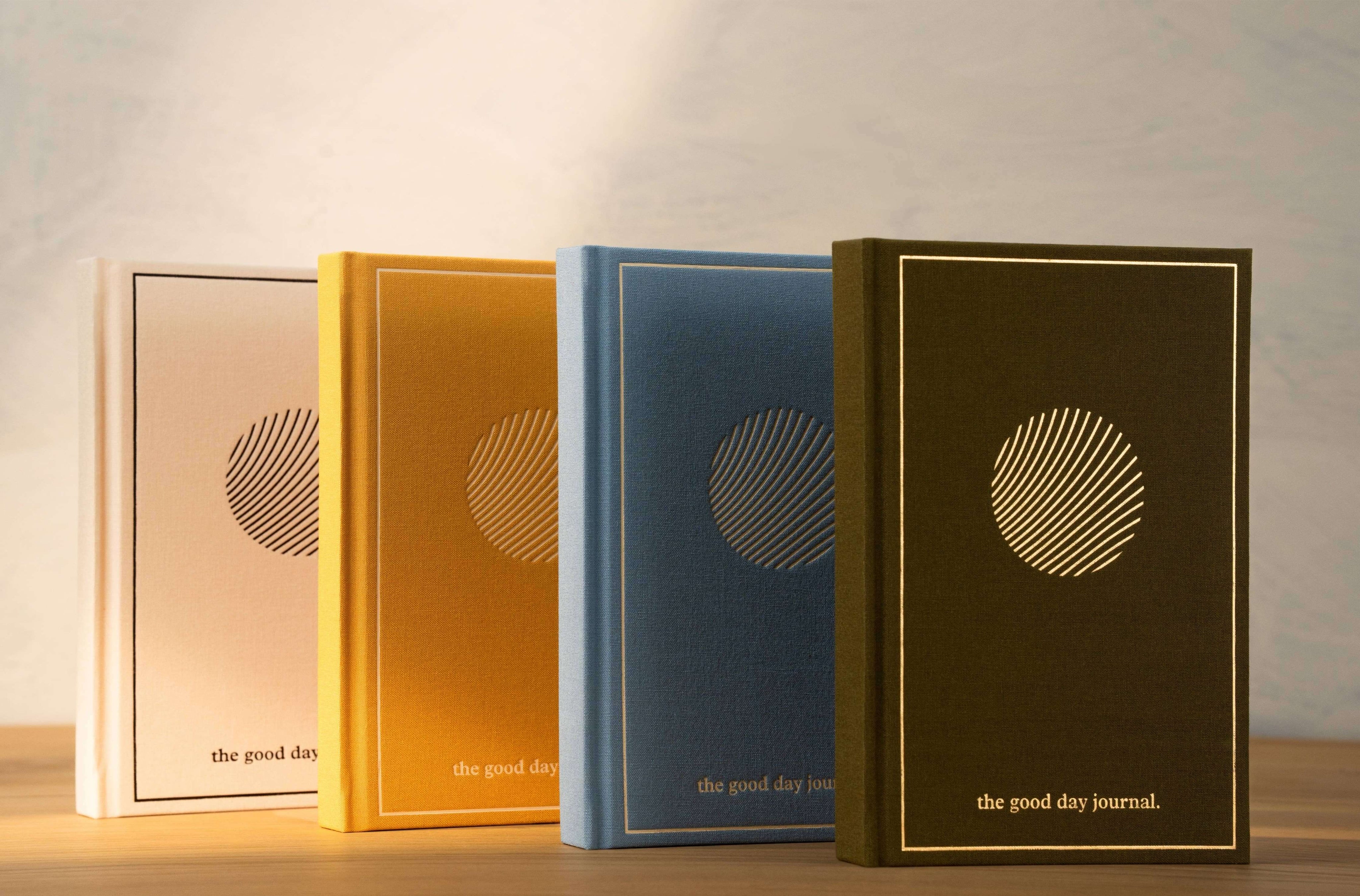 Four 'the good day journal' books in different colors on a wooden surface with a light background.