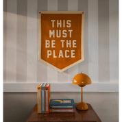 Decorative banner with 'THIS MUST BE THE PLACE' text on a wall above books and a lamp.
