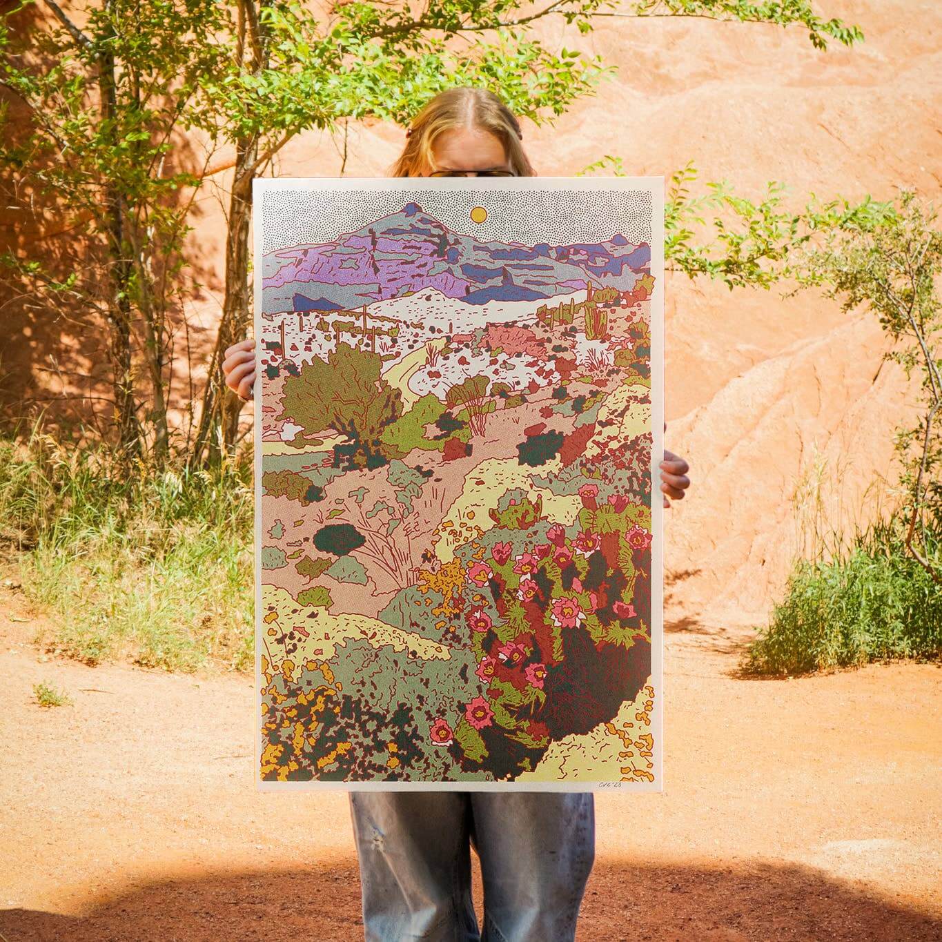 Person holding a colorful landscape illustration in front of a natural rock formation.