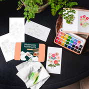 Watercolor painting kit with watercolor paper, paint set, and a plant on a dark surface.